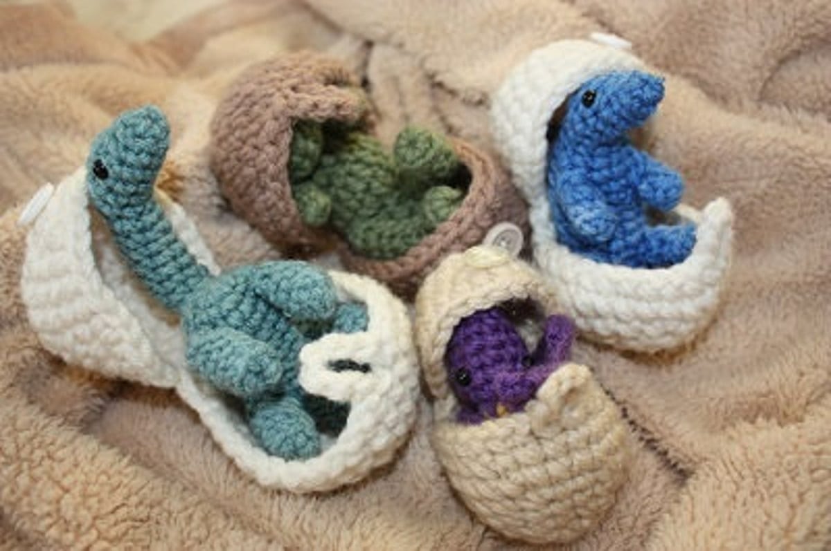 Small stuffed blue, purple, and green baby dinosaurs hatching from white and light brown crochet eggs on a beige blanket.