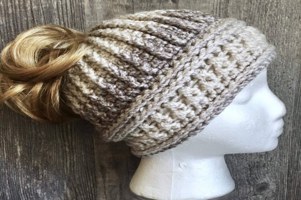  A white mannequin wearing a cream and gray ombre style hat with a messy bun pulled through the top on a wooden background.