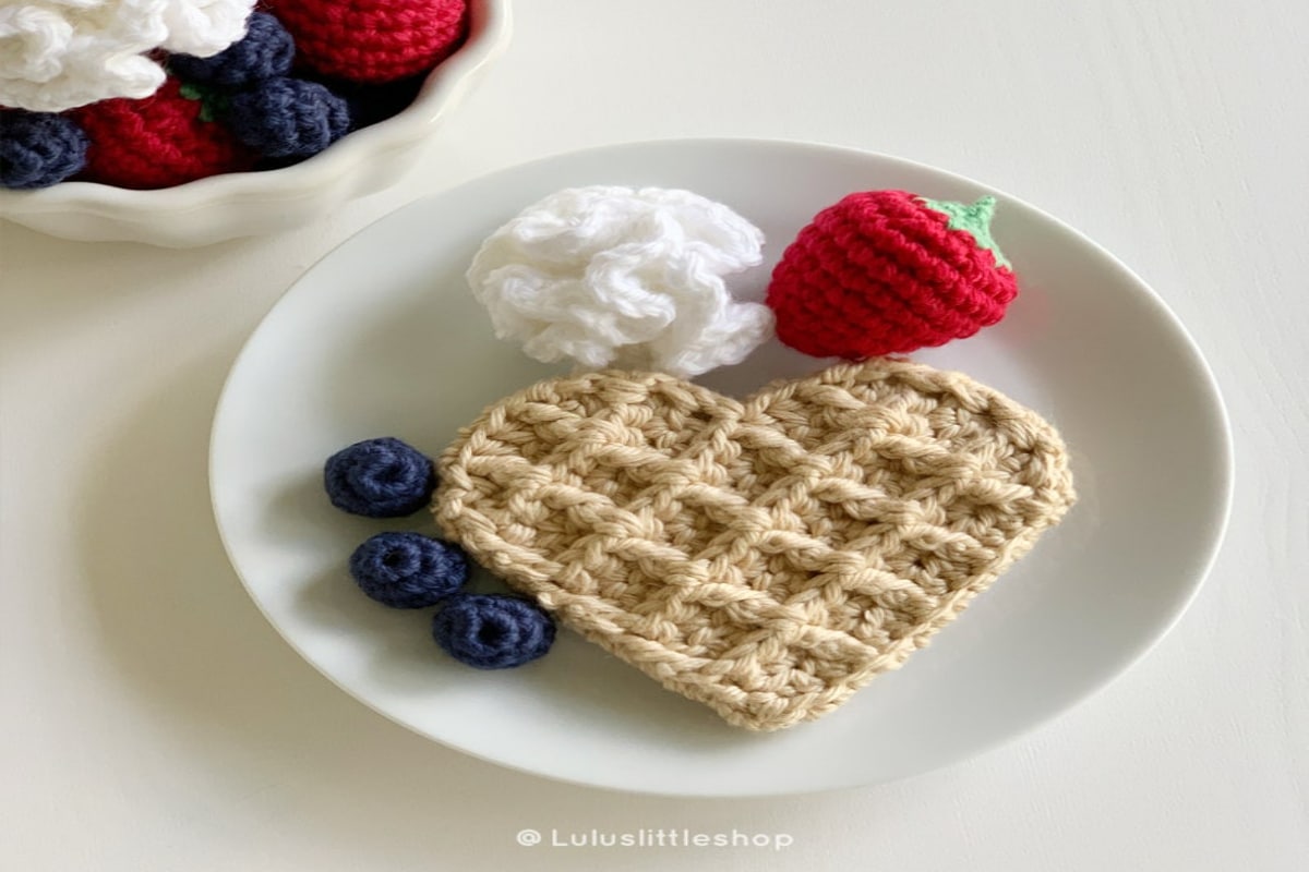 Beige waffle stitch crochet shaped as a heart-shaped waffle on a plate next to crochet blueberries, a strawberry, and whipped cream.
