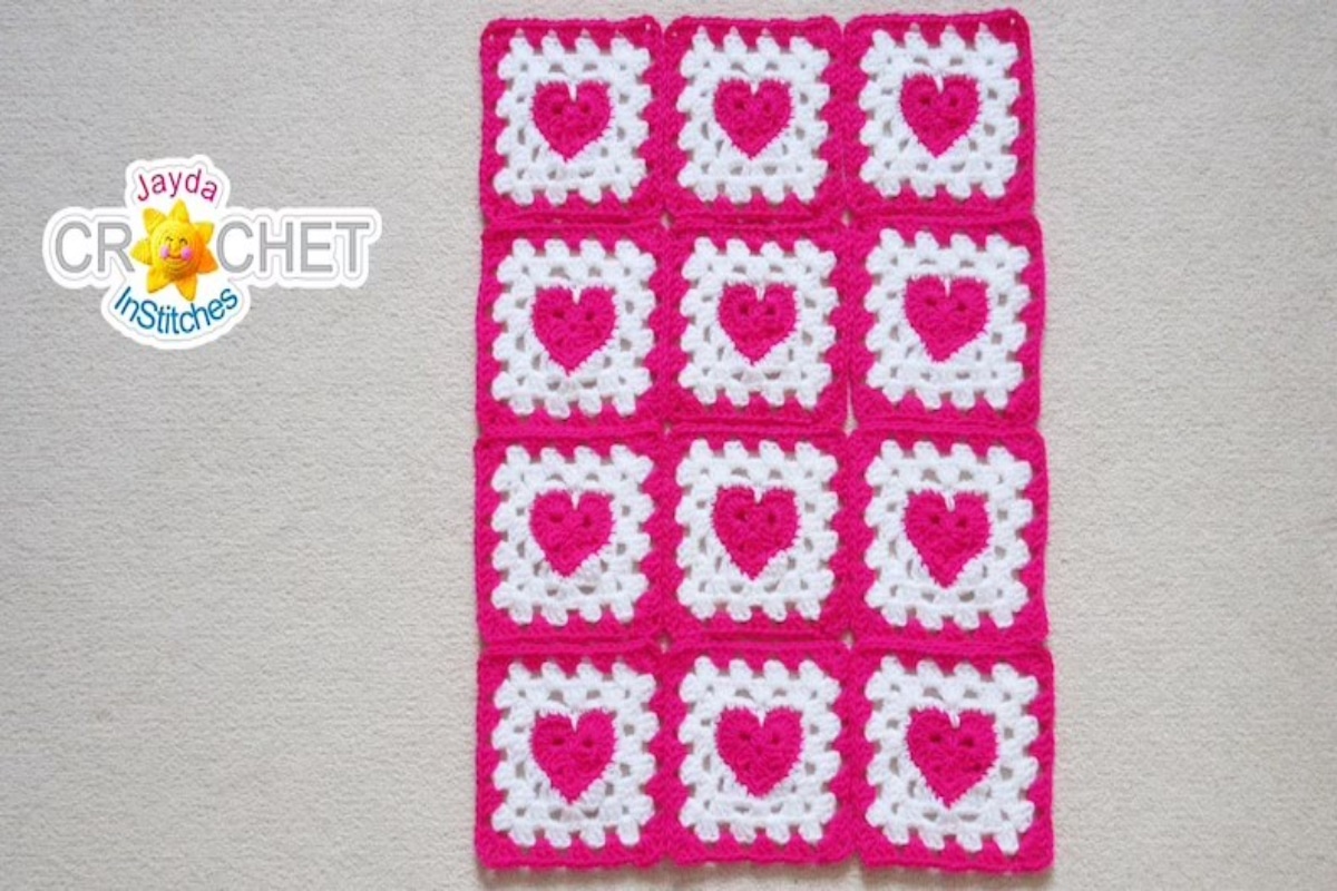 Pink crochet blanket with 12 white squares each with a pink heart stitched into displayed on a white background.