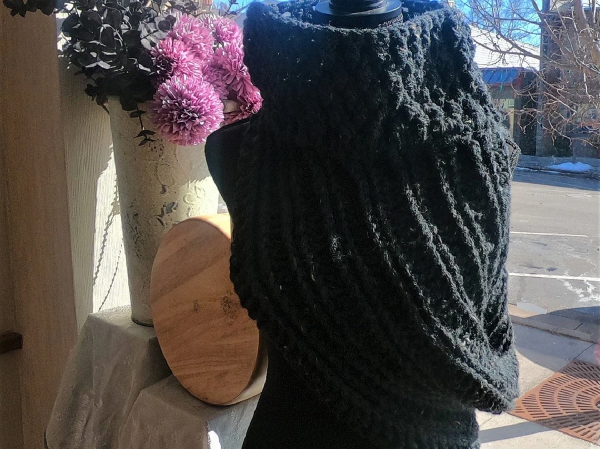 A dark green crochet cowl with a high neck on a mannequin next to a vase of flowers and a window.