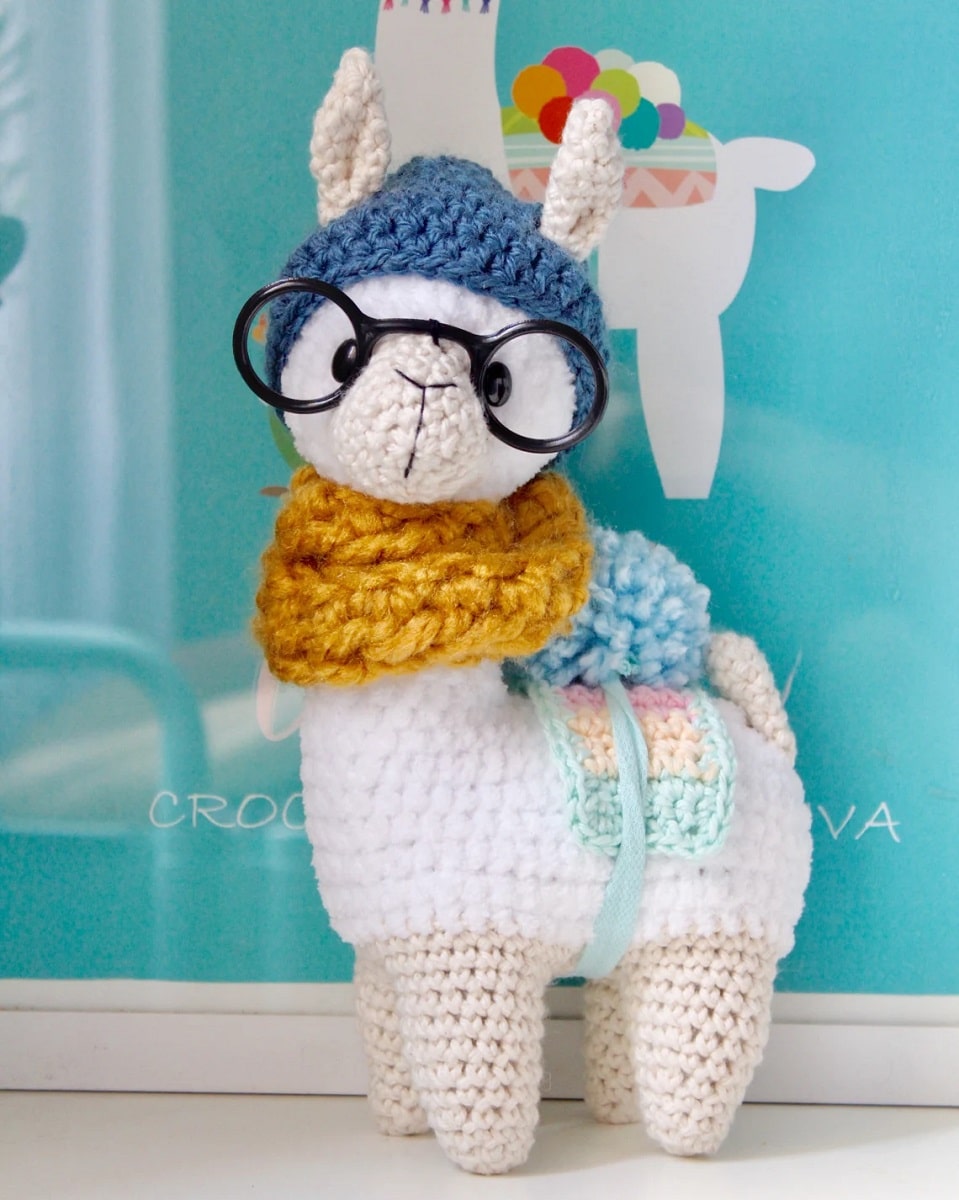 White stuffed crochet llama with a blue bobble on its middle, a chunky mustard yellow scarf, black glasses and a dark blue beanie on.