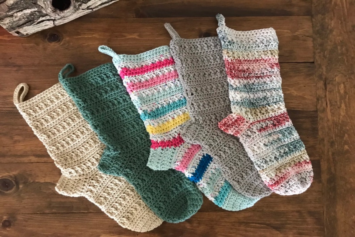 Cream, green, striped, and gray stockings on a wooden floor. Stockings all feature loops to attach and a chunky knitted design. 