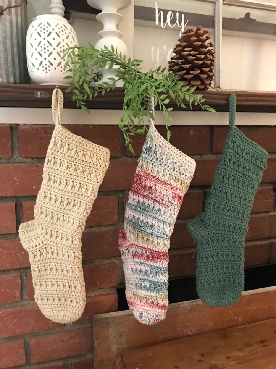 A cream crochet Christmas stocking hanging next to a cream, red, blue, and yellow striped stocking and a green stocking.