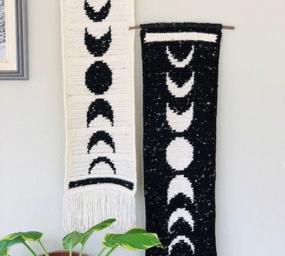 A white crochet wall hanging with black half moons, crescent moons, and full moons running down the center next to the same wall hanging in black.