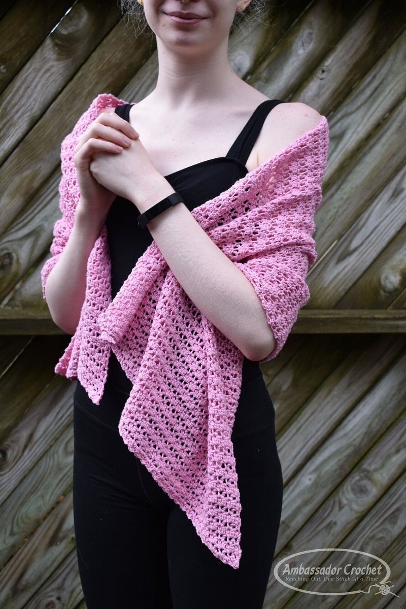 Woman wearing a pink crochet prayer shawl wrapped around her shoulders and arms and a black jumpsuit.