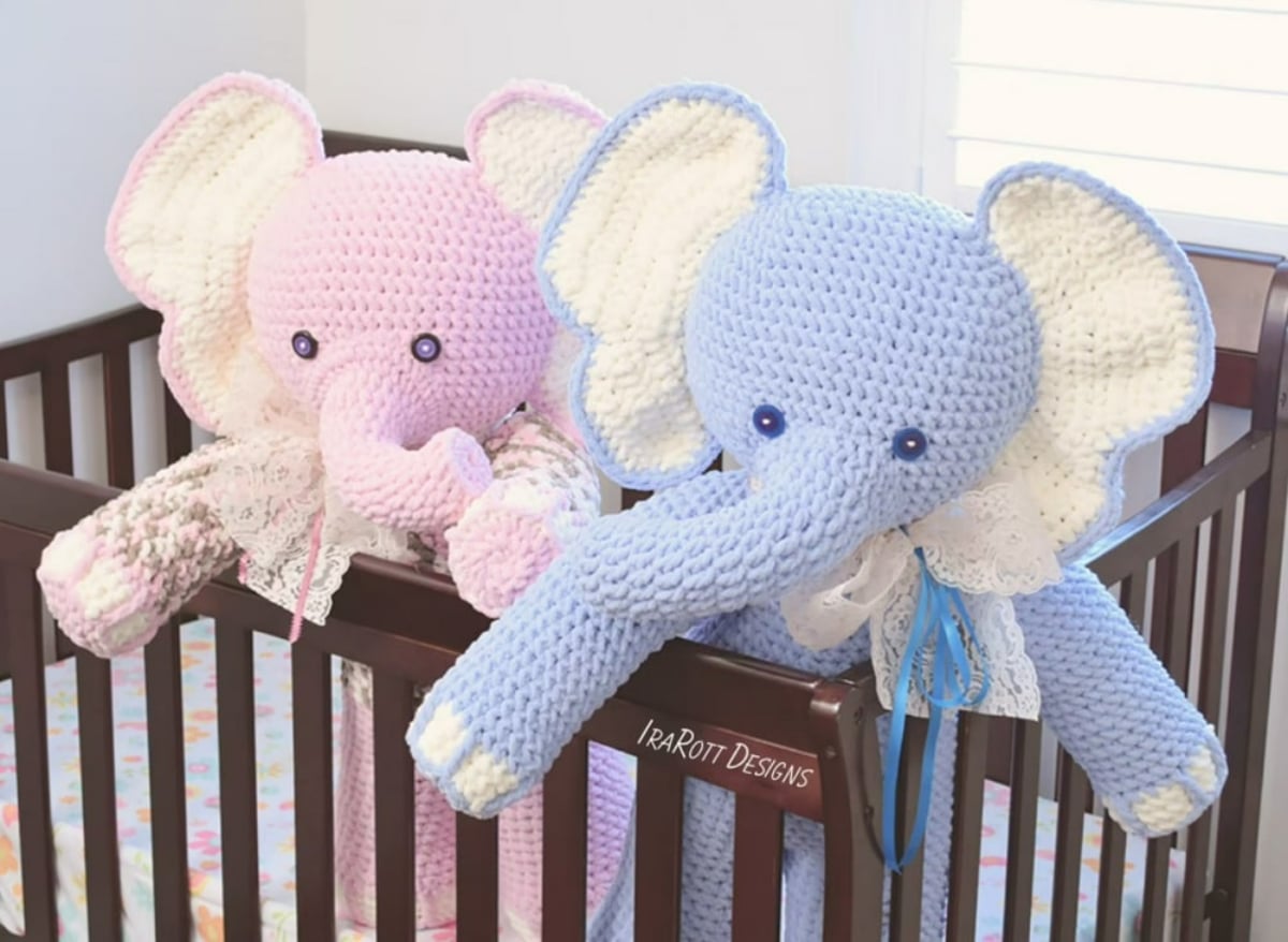 A large blue crochet elephant with large yellow ears standing in a crib next to a large pink elephant with yellow ears.