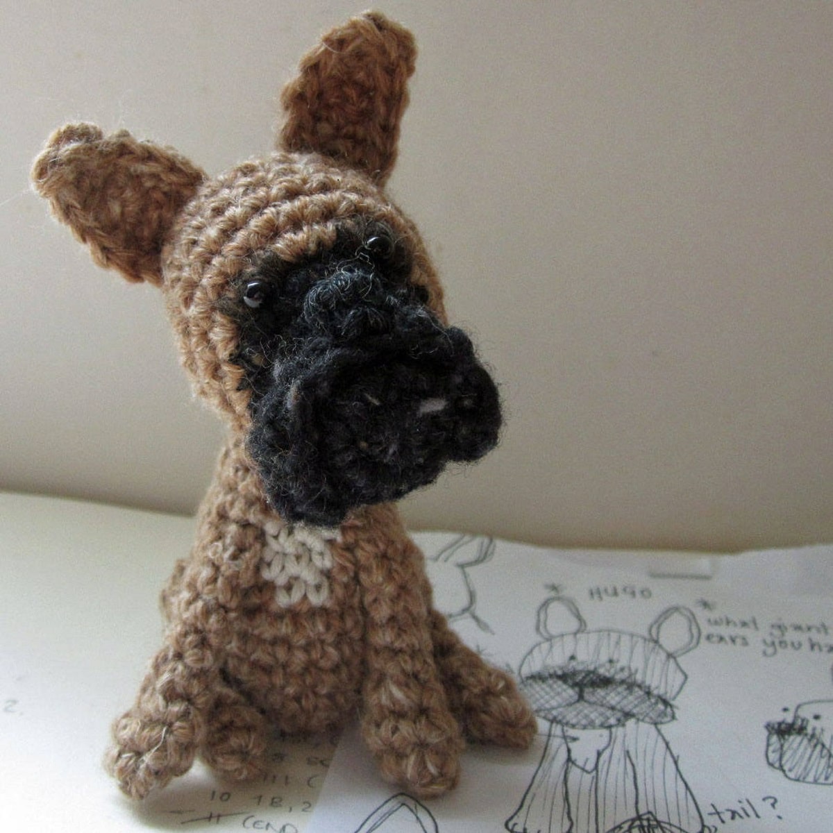 Small crochet stuffed French Bulldog with a brown body and ears and a black face sitting on a white background.