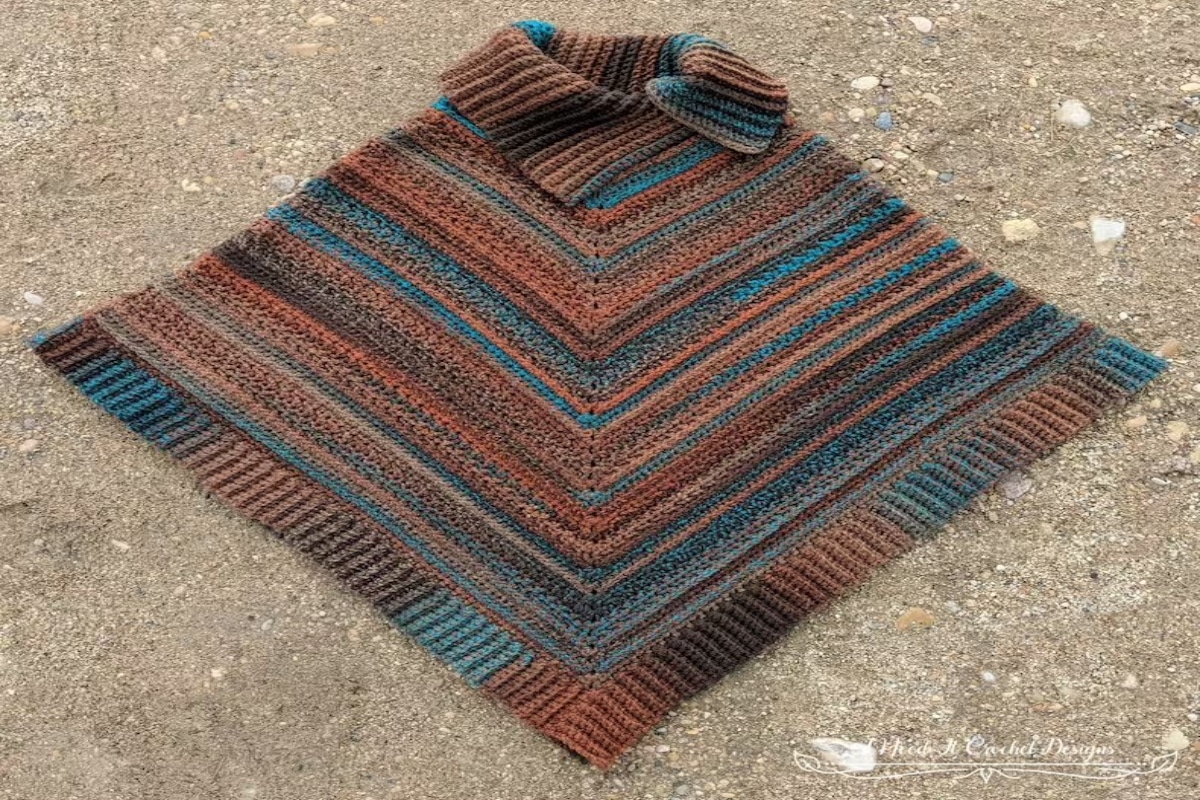 A crochet poncho using blue, brown, and orange yarn stitched in diagonal stripes with a vertical striped trim laid out on a sandy background.