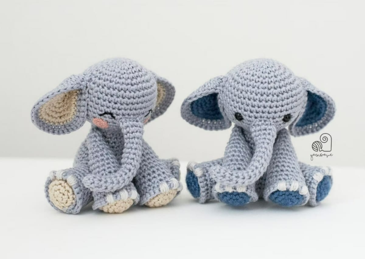 Two crochet gray elephants, one with dark blue feet and ears, the other with beige feet and ears, sitting next to each other. 