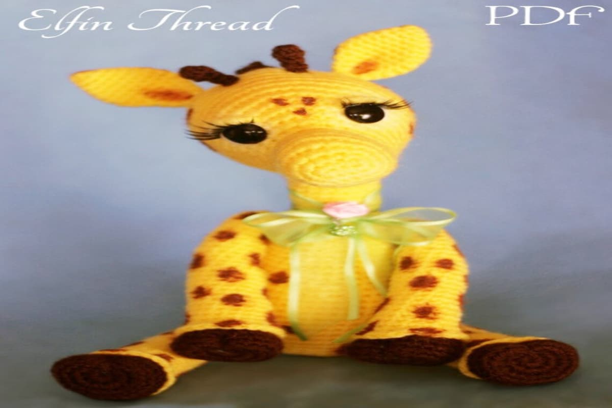 Large crochet giraffe with orange circles on its arms and legs, sitting down with a bow around its neck, black eyes, eyelashes, and small brown circles on its face.