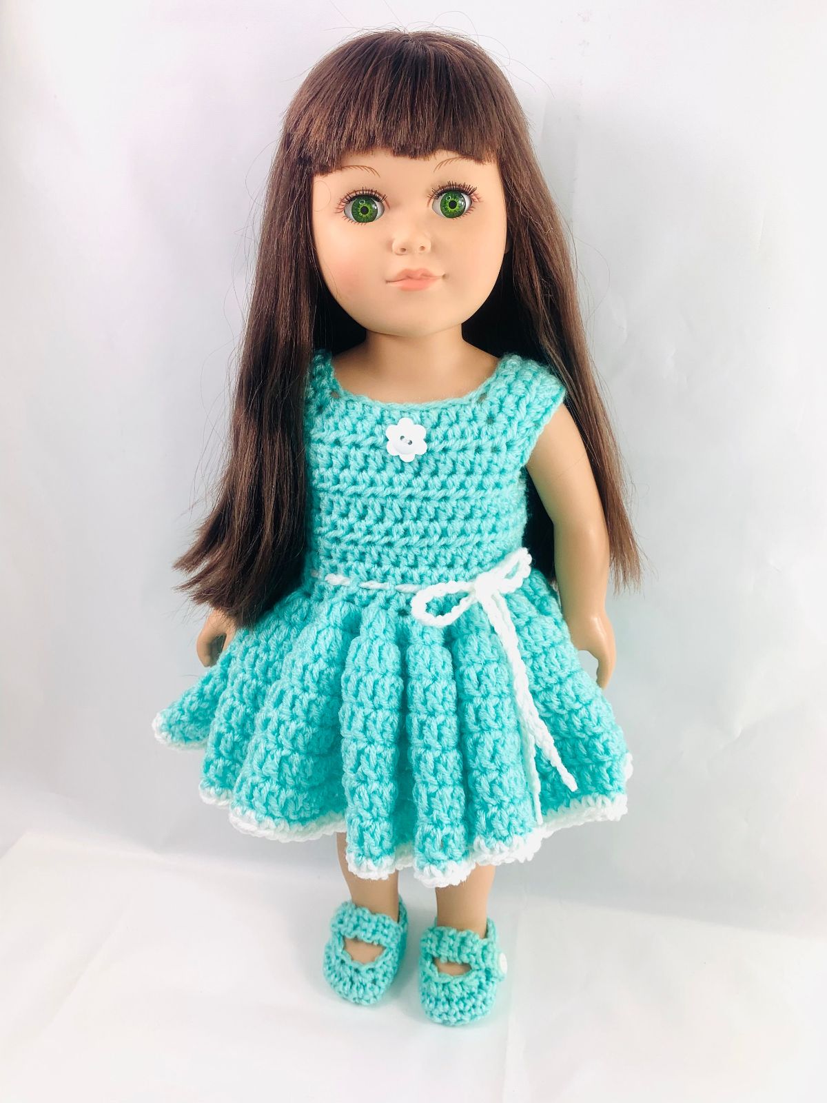 Brunette doll wearing a sleeveless blue crochet dress with a white bow around the waist and matching blue slip on shoes.