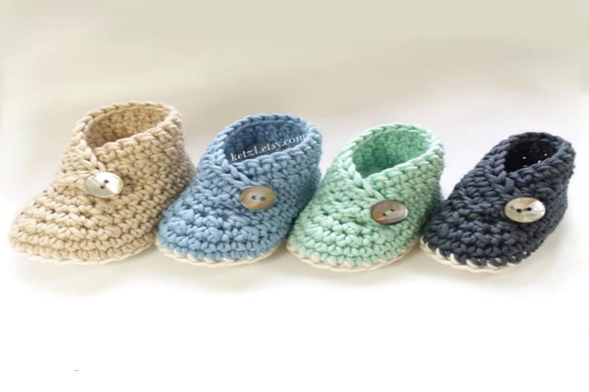 Four kimono style crochet baby booties with a button to secure. Booties are black, mint green, blue, and yellow with cream soles.