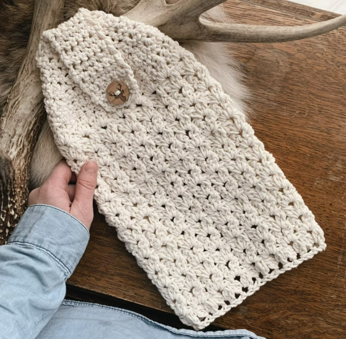 A white crochet hand towel with a pointed end and a flap with a small button to secure on a dark wooden table.