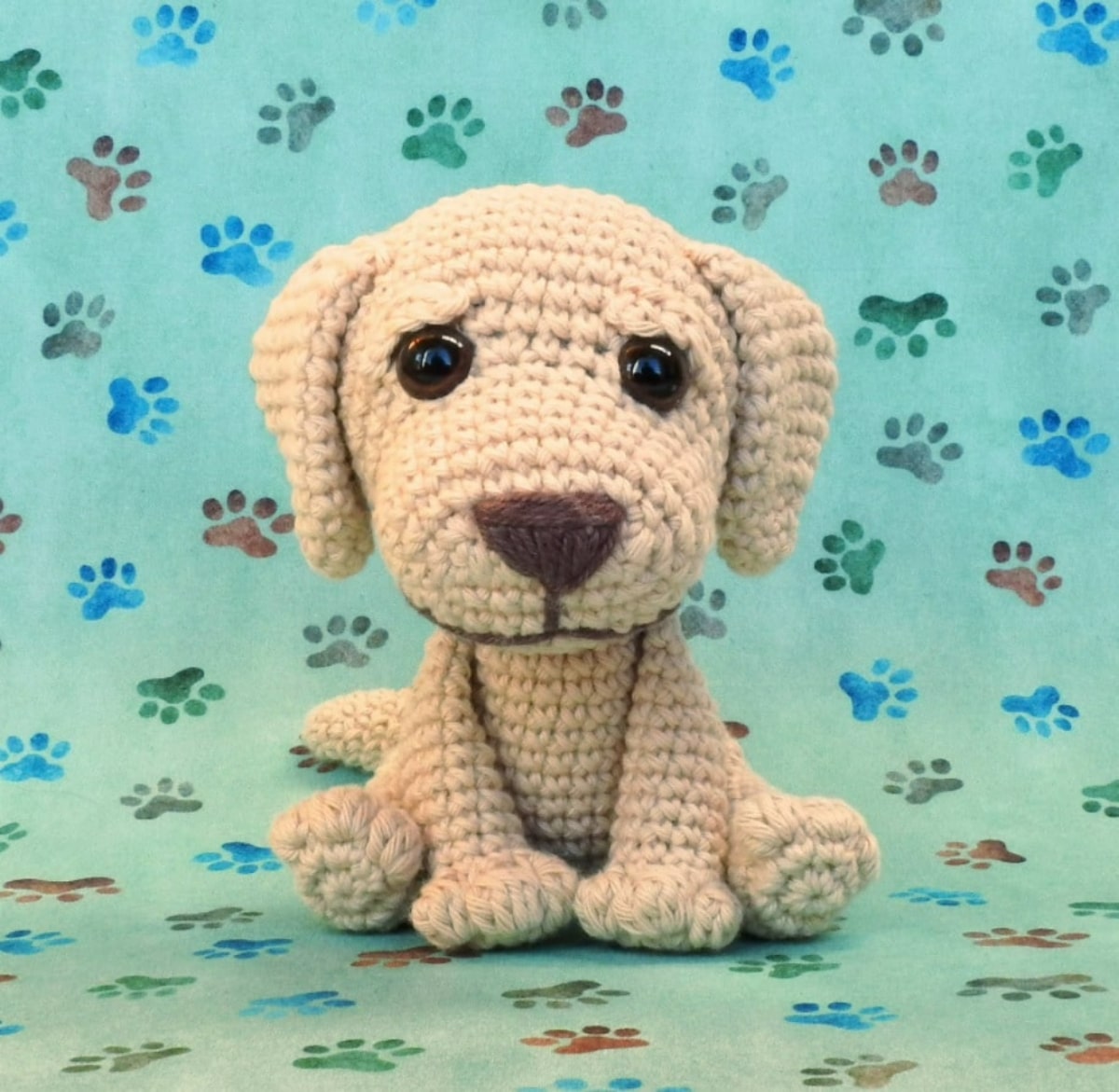 Small crochet stuffed Labrador with an oversized head, and large orange nose sitting on a green background covered in paw prints.