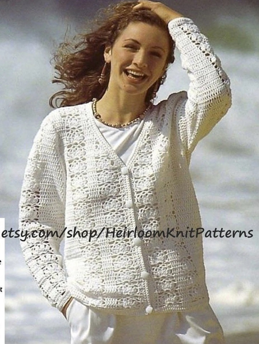 Brunette woman on a beach wearing a white lacy crochet cardigan with long sleeves buttoned up over a white t-shirt.