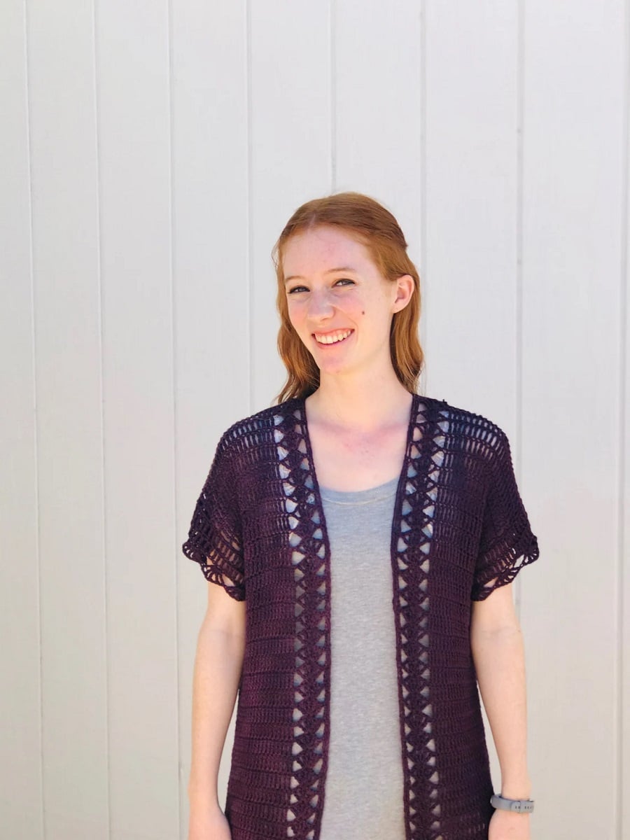 Redhead woman wearing a purple short sleeves lacy crochet cardigan over a gray vest.
