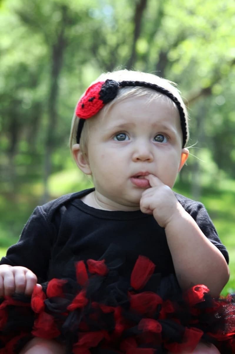 Blond baby wearing a slim black crochet headband with a red and blag lady bug on the left hand side with black spots on.