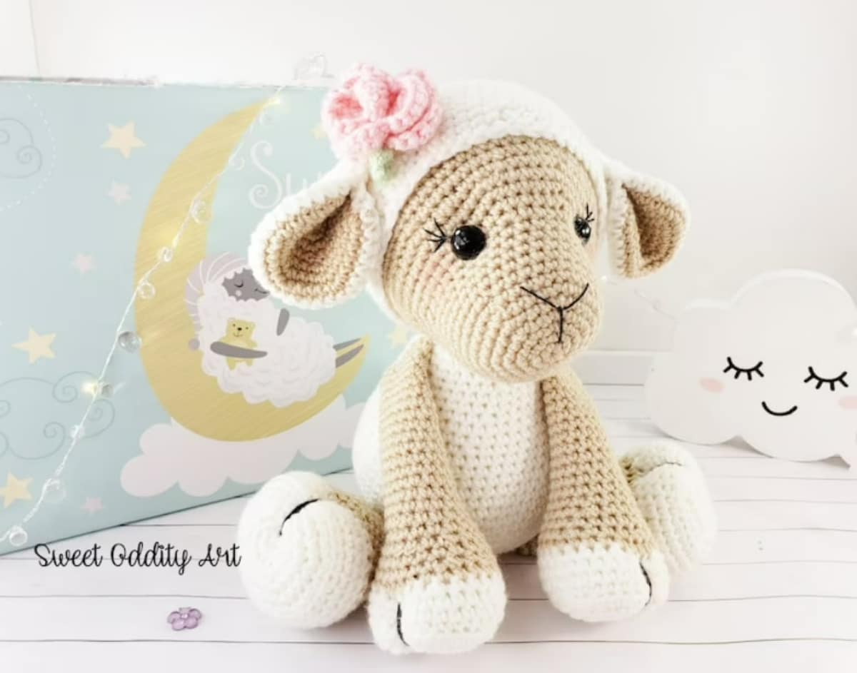 White stuffed crochet lamb with a light brown face and legs sitting on a white wooden floor with a pink flower on its head.