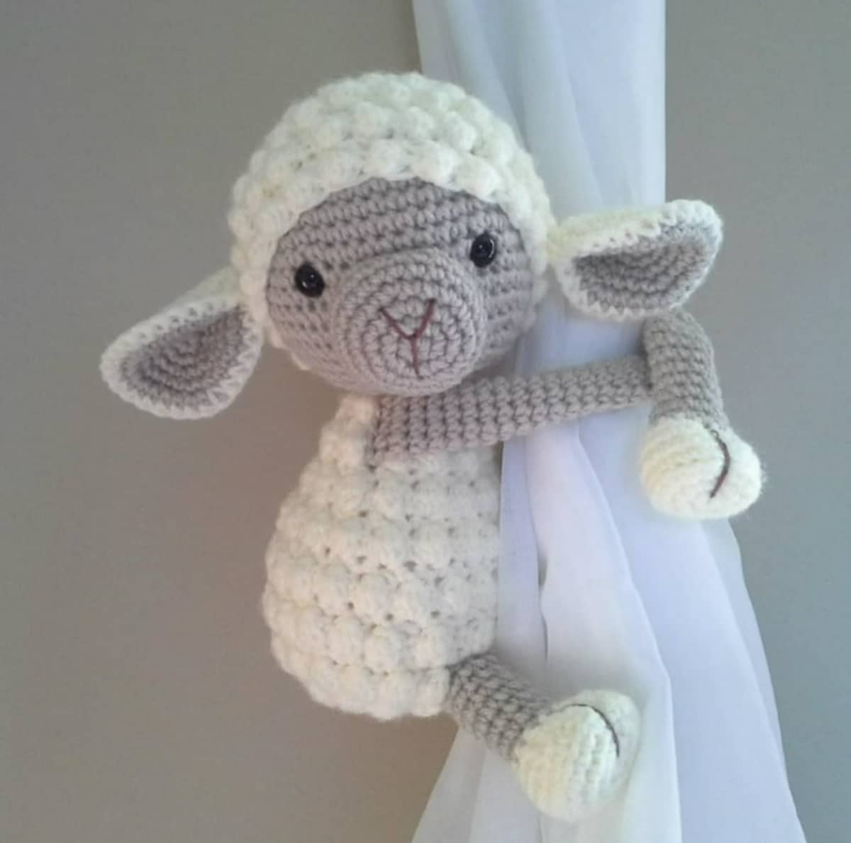 White crochet lamb with a textured body and head and a brown face and legs wrapped around a white curtain.
