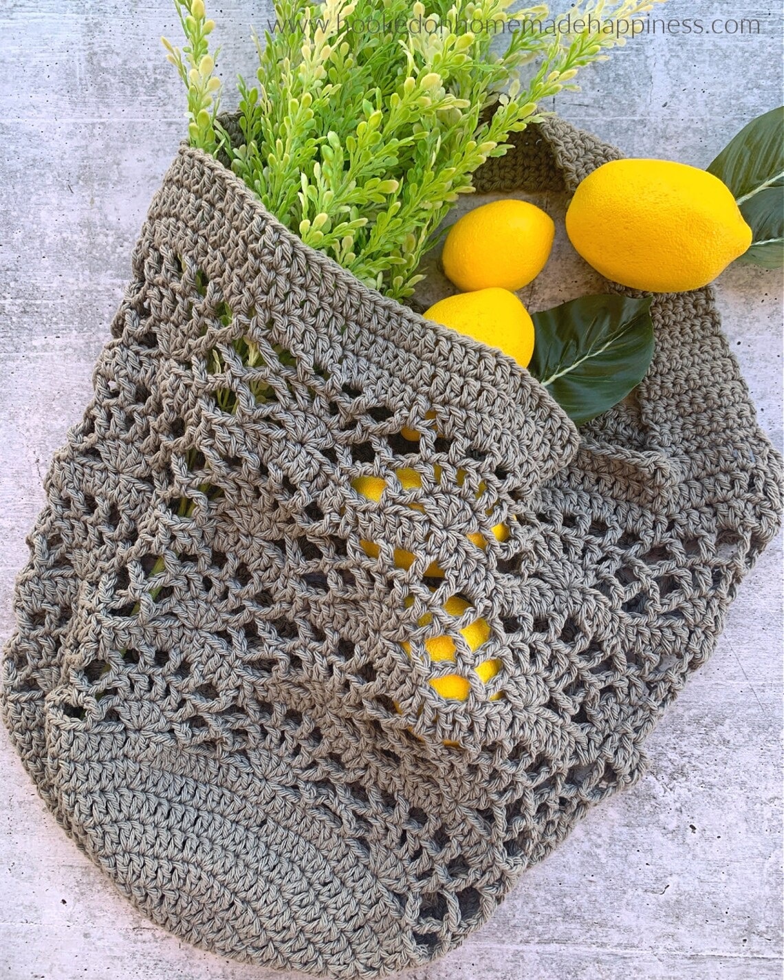 A light gray lattice style crochet market bag with a fan shaped pattern on a cream background with lemons and leaves in the bag.