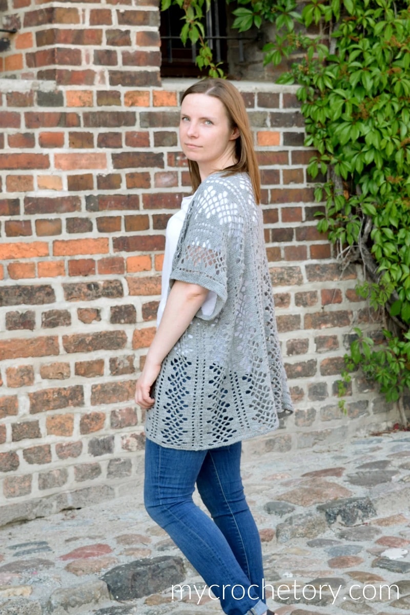 Brunette woman standing sideways wearing a gray lacy crochet cardigan with short sleeves and a diamond pattern all over.