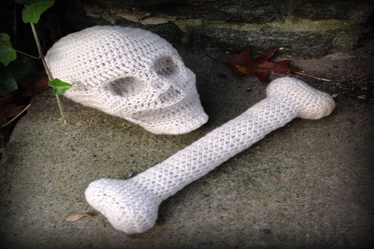 A large white crochet skull with a large bone in front of it on a concrete floor outside.