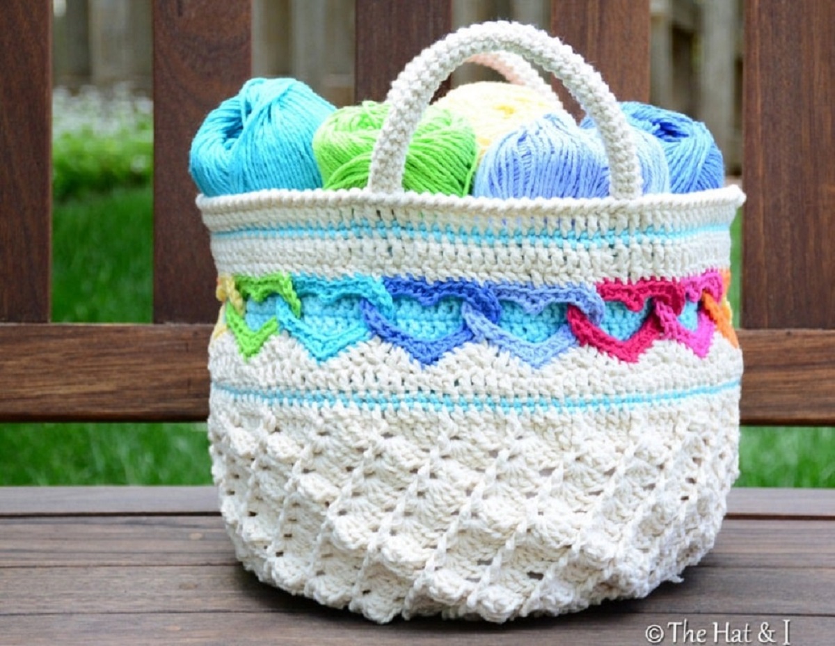 Cream crochet bag with colorful linked hearts stitched around the middle of the bag, with thick cream straps and blue and green yarn inside the bag.