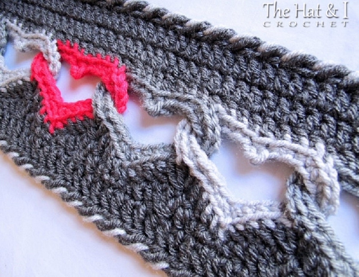 Gray crochet scarf with a braided trim and red, gray, and white linked hearts running down the middle of the scarf.