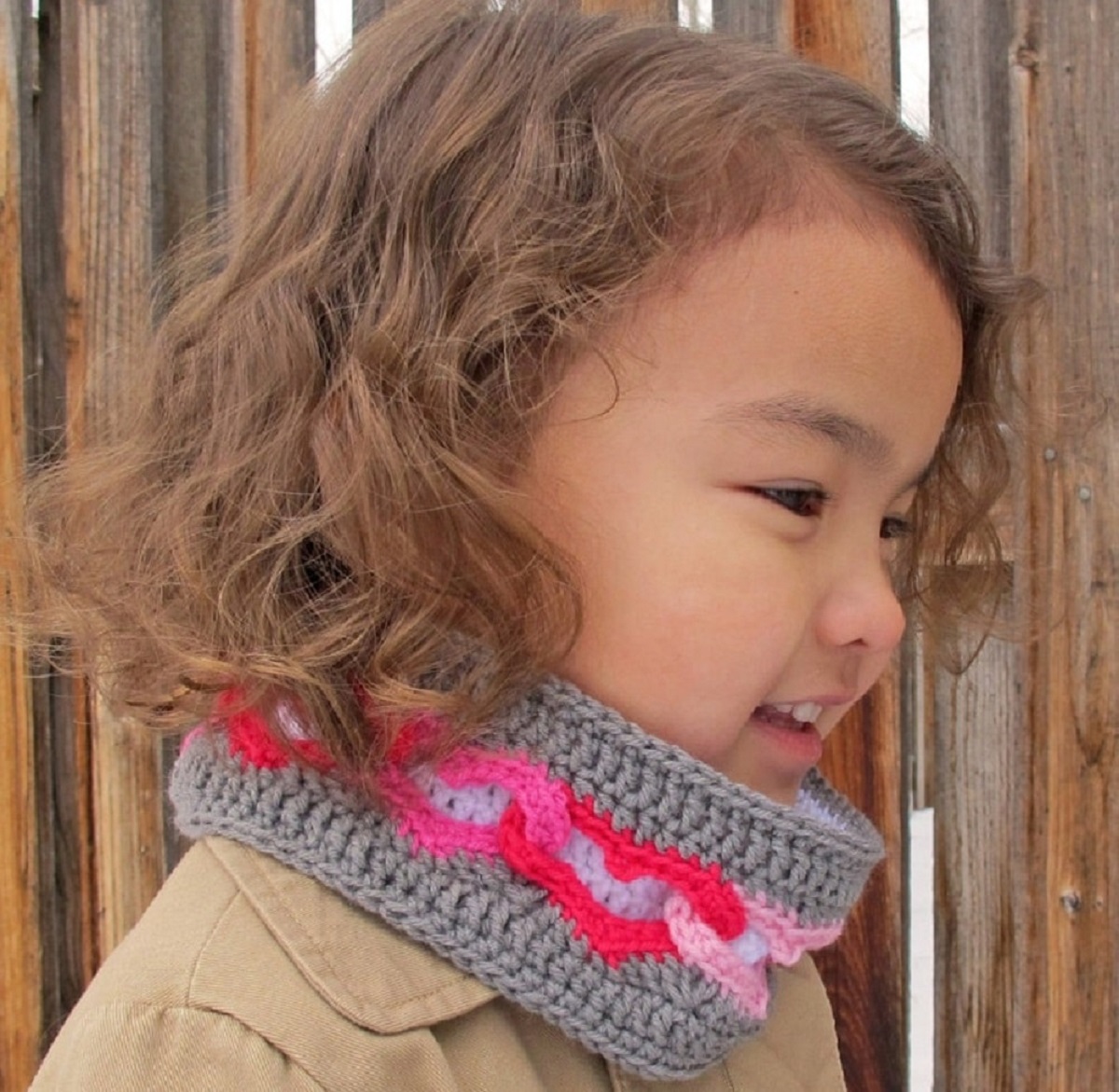 Small child facing sideways with a gray crochet neckwarmer with bright pink and pale pink linked hearts around the middle of the warmer.