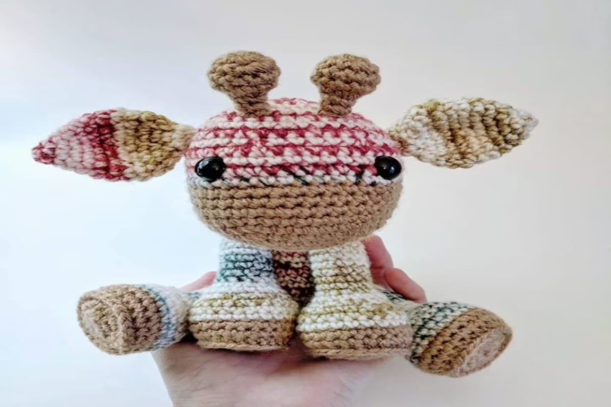 Alpine style multicolored crochet giraffe with pink, blue, and brown stripes across its face and body on a white background.