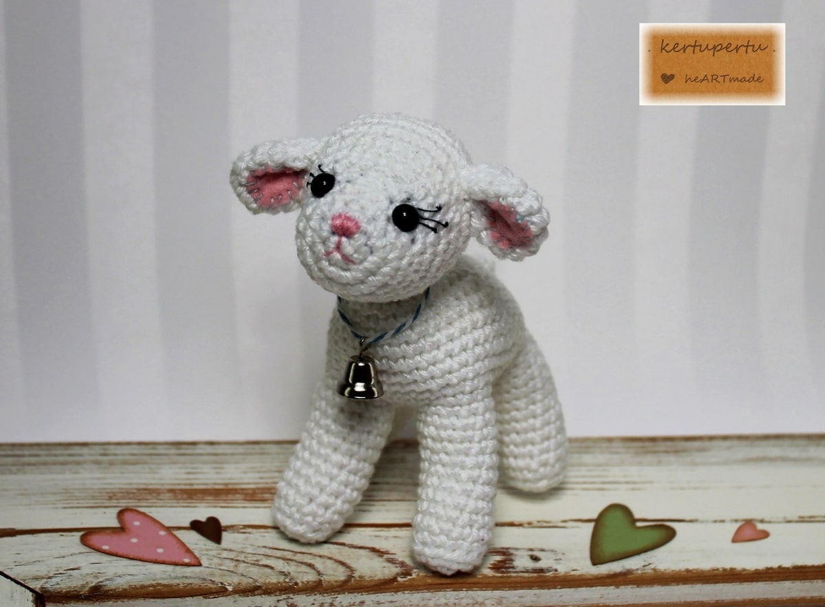 Small white crochet lamb with pink ears and a bell around its neck standing on a wooden floor.