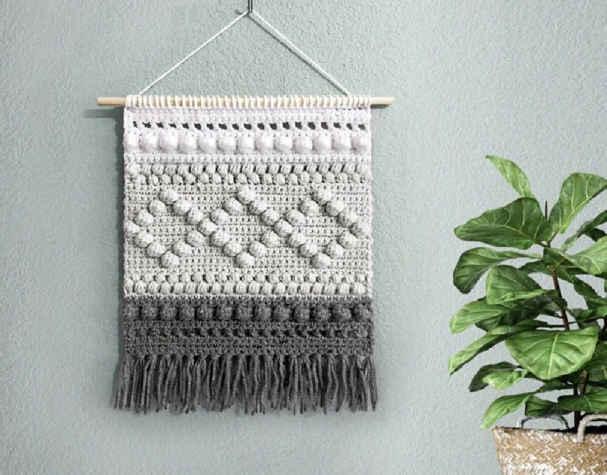 A cream, beige, and gray striped crochet wall hanging with gray tassels at the bottom, and a diamond pattern in the center.