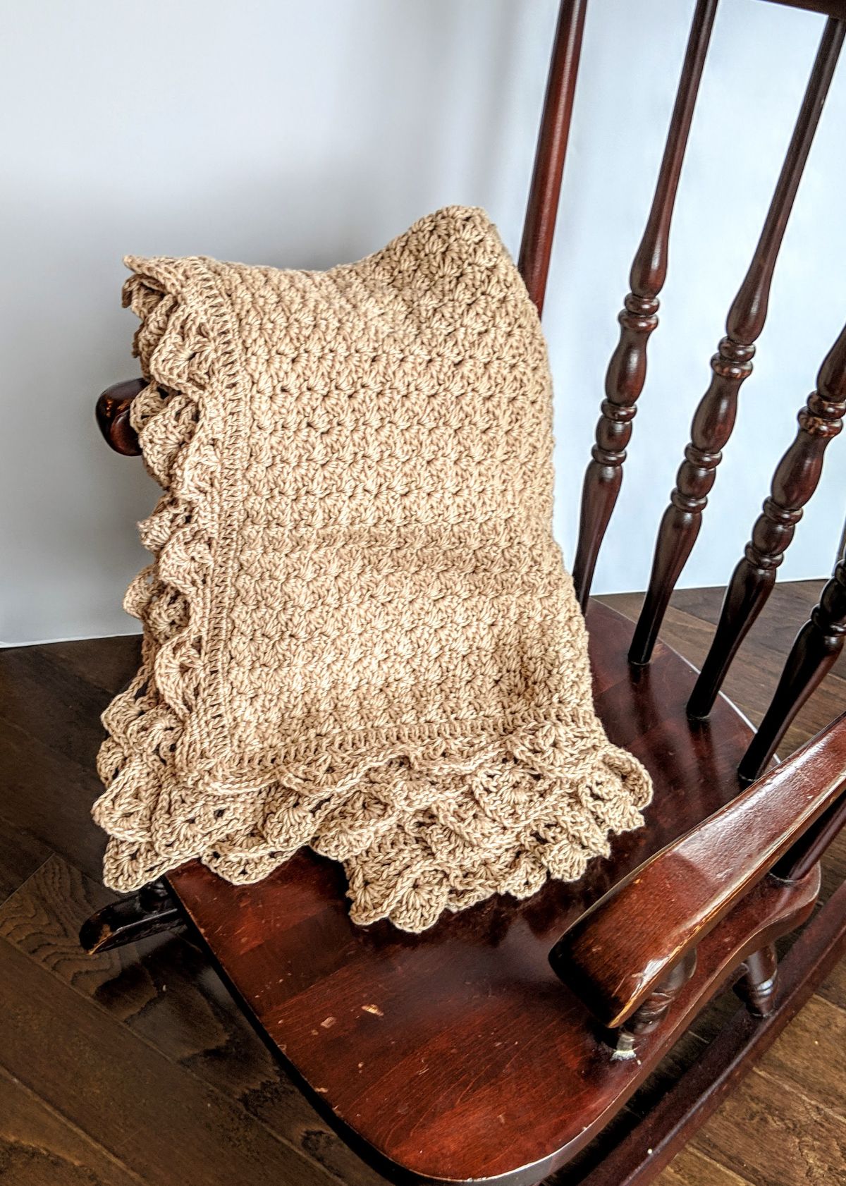 A light brown crochet baby blanket with a long lace trim folded into a rectangle over a dark wooden rocking chair.