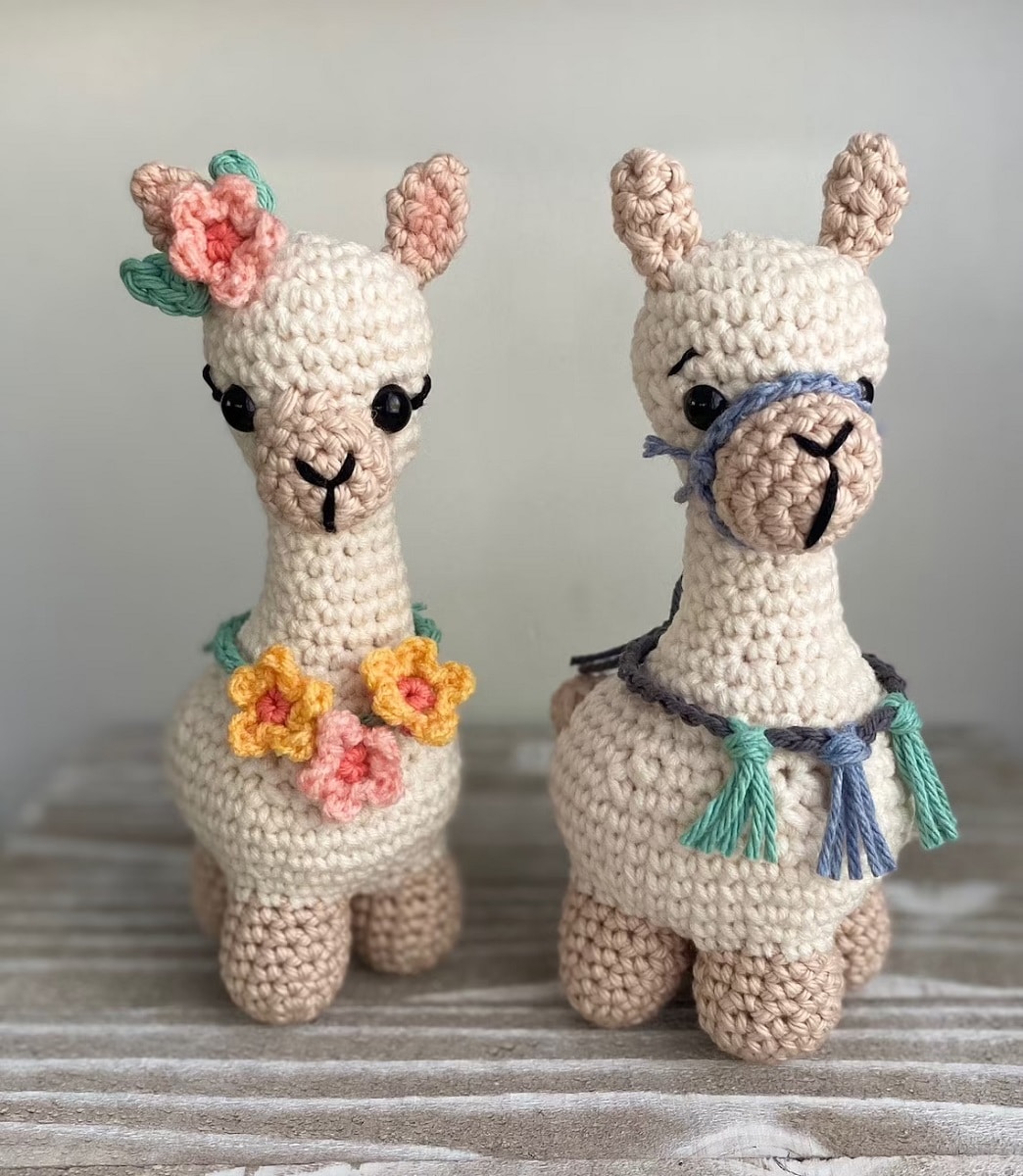 Two cream crochet llamas with light brown ears and legs. One llama has orange and pink flowers around its neck and the other blue and green tassels.