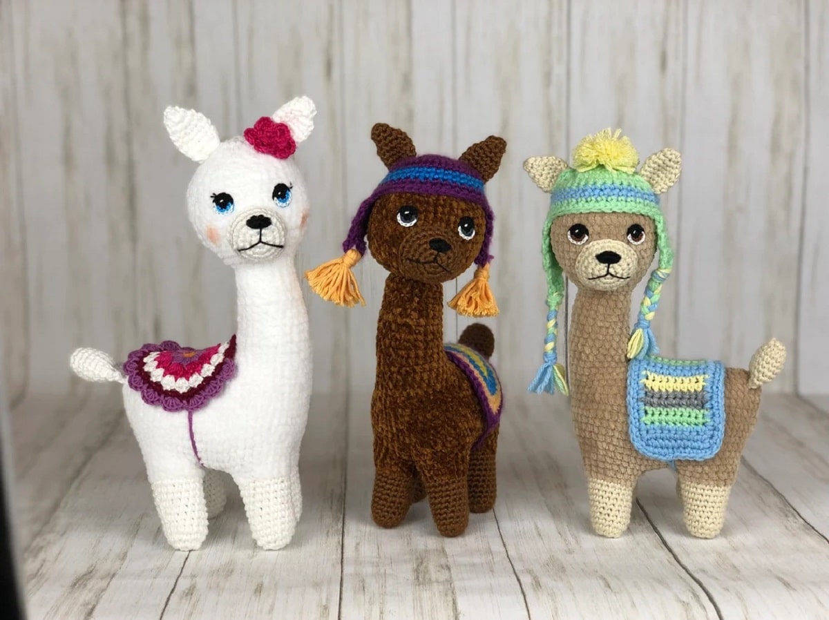  White, light brown, and dark brown crochet llamas standing next to each other on a wooden background wearing red and blue seats on their backs.