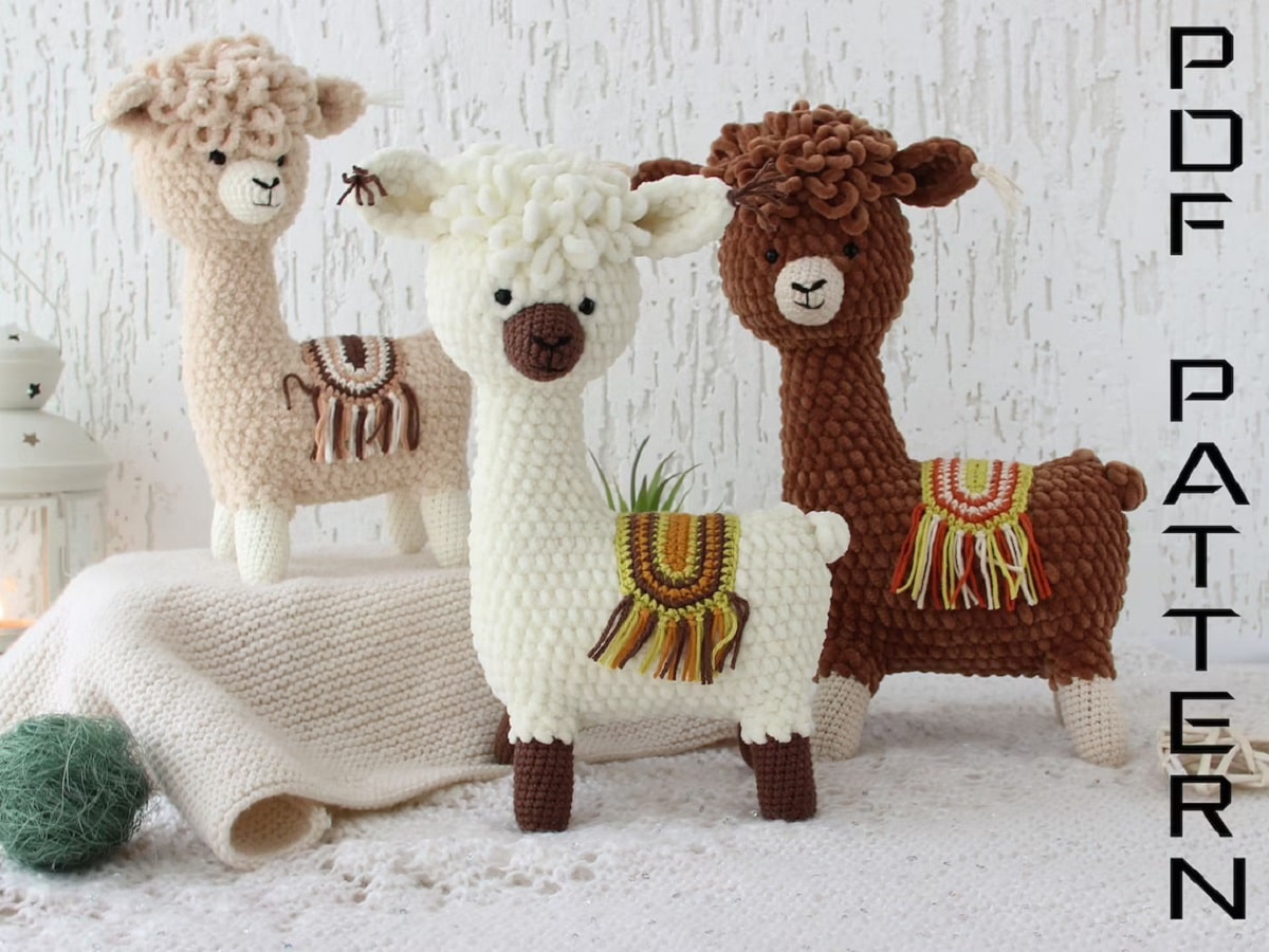 Cream, white, and brown stuffed crochet llamas with multi-colored seats with tassels on their middles standing on a white background.