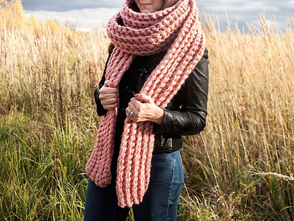 Woman standing outside wearing a chunky, oversized pink crochet scarf wrapped around her neck.