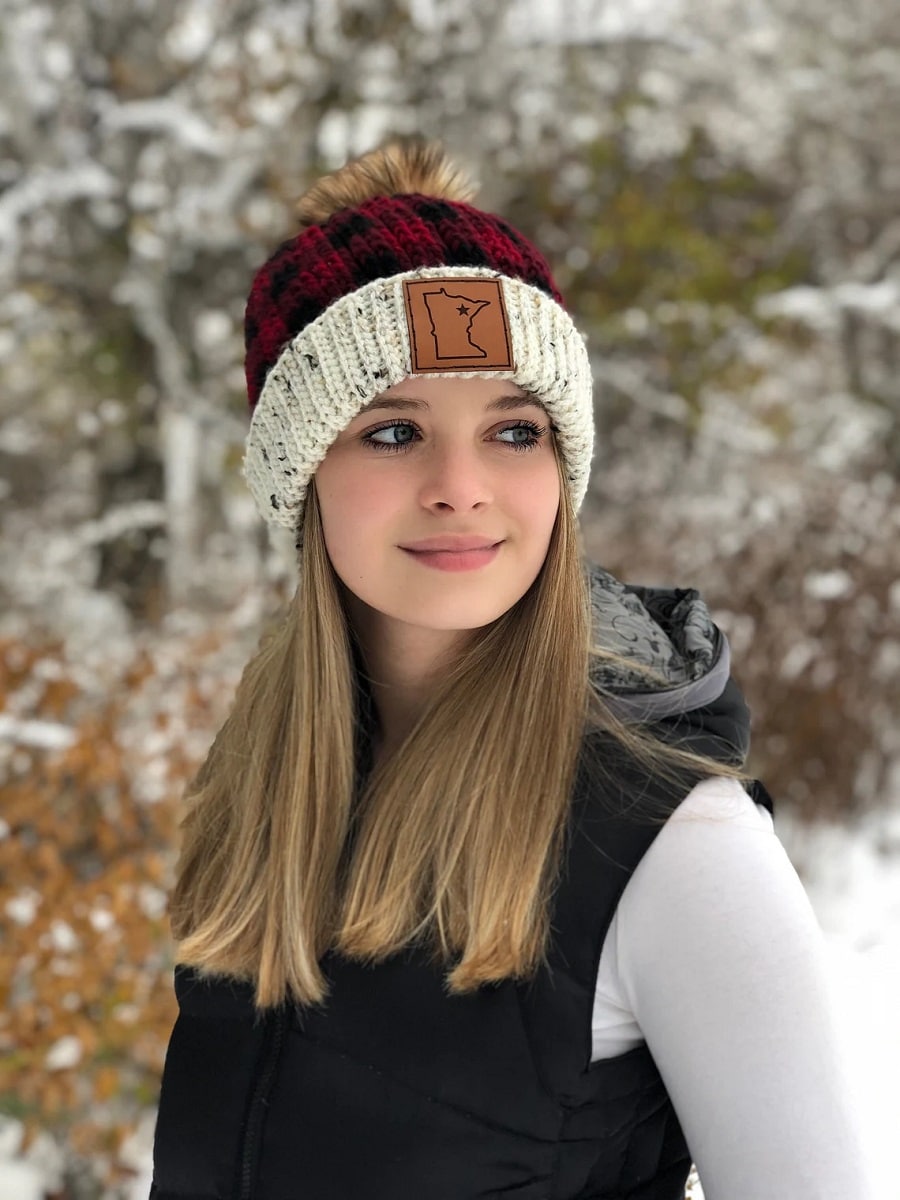 Blonde woman standing sideways in the snow wearing a black and red plaid crochet beanie hat with a thick white banding around the bottom.