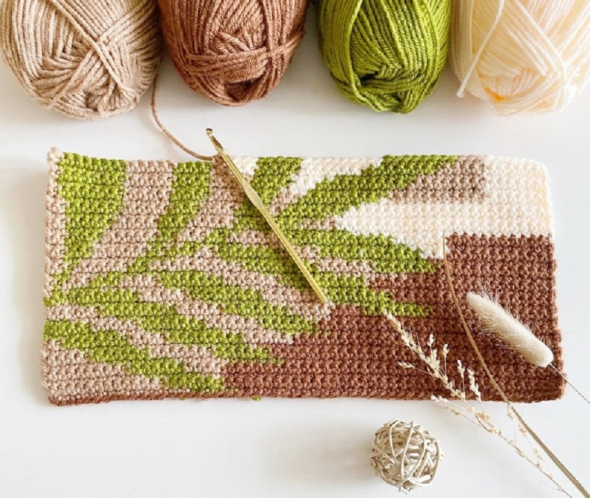 A brown crochet rectangle with a green leaf stitched on top next to balls of yarn and a crochet hook on top.
