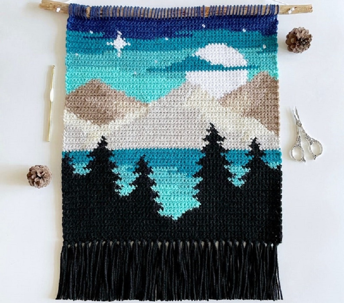 Crochet wall hanging of beige and brown mountains, a blue sky with stars, blue lake and black tree outlines. 