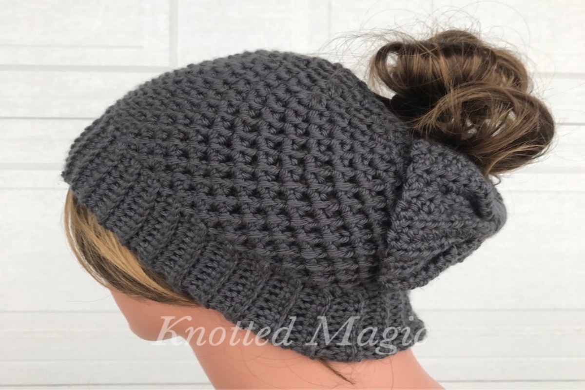 A brunette mannequin wearing a slouchy gray crochet beanie with a messy bun pulled through the top.