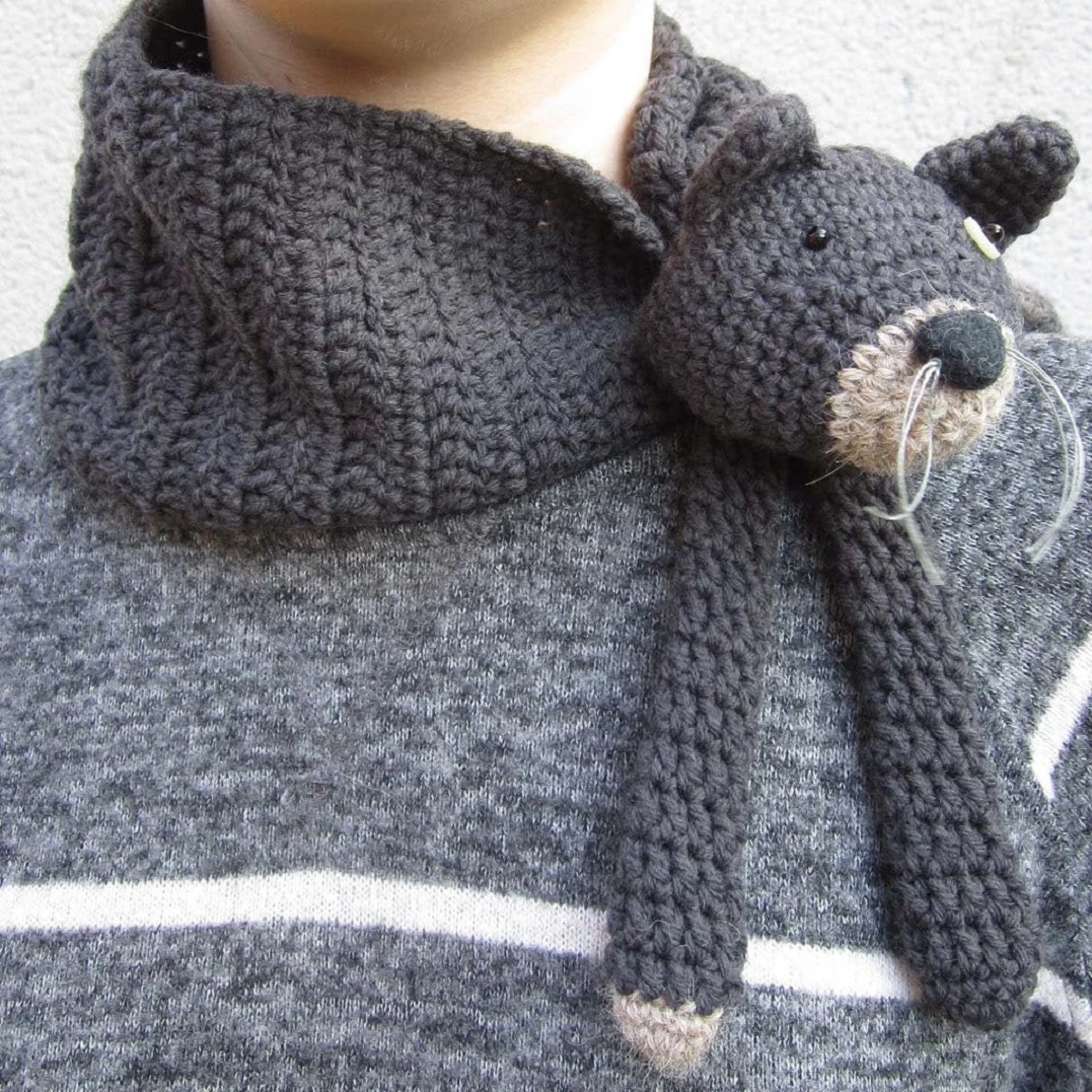 A woman wearing a crochet scarf with a stuffed black cat’s face and front legs attached to the right side.