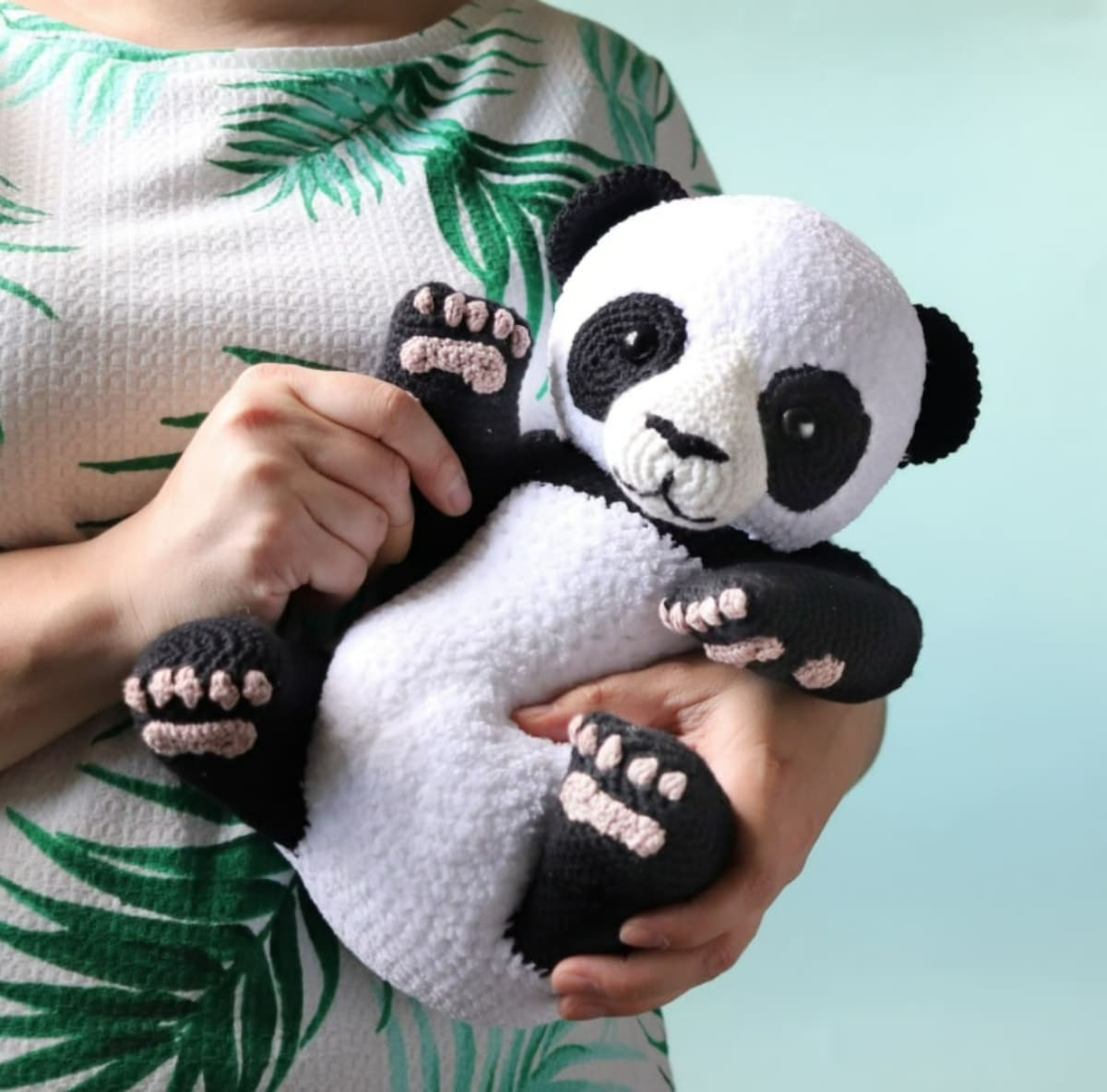 Large crochet and stuffed black and white panda with pink paws and feet being held like a bay by a woman in a pink and green top.