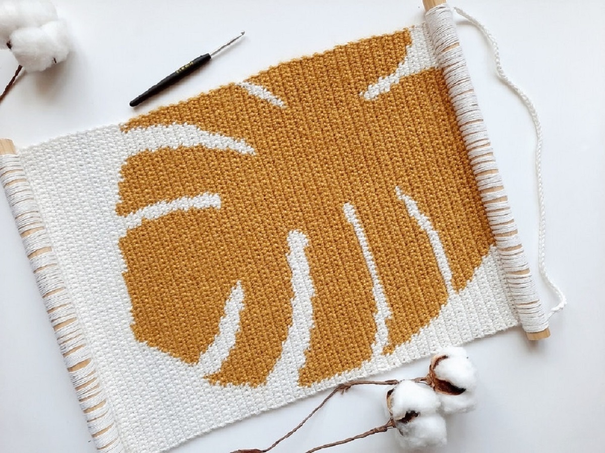 A white crochet wall hanging with small fringing on the bottom on a large brown leaf on the center.