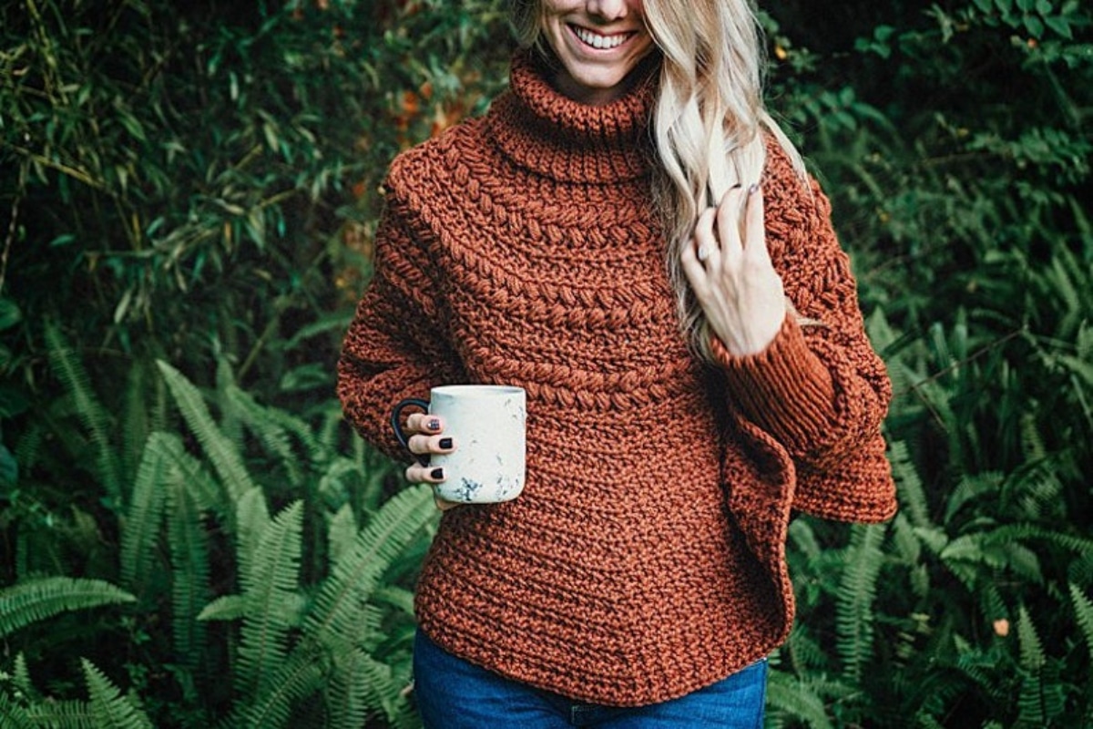  A smiling blond woman holds a marble-colored cup, wearing a long-sleeved rust-colored poncho with a high neck.