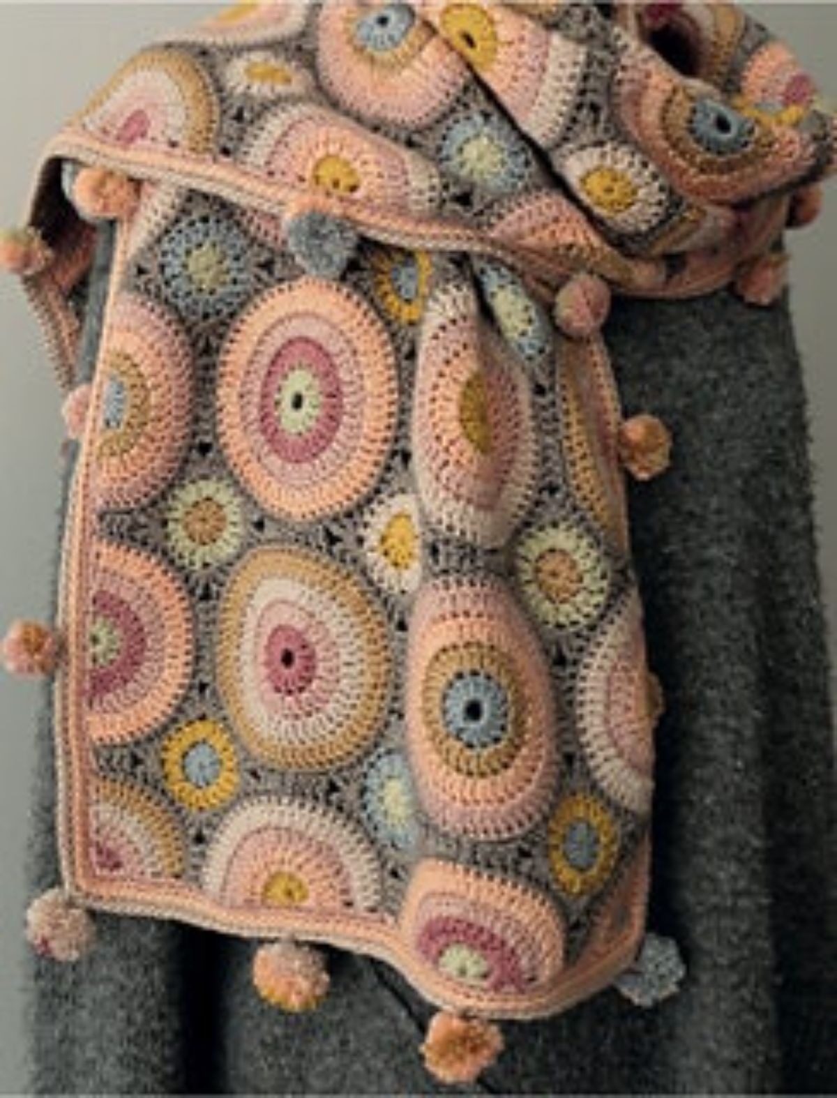Gray scarf featuring pink, blue, yellow, and gray crochet circles all over with a pale pink trim and pink and gray pom poms along the edges.