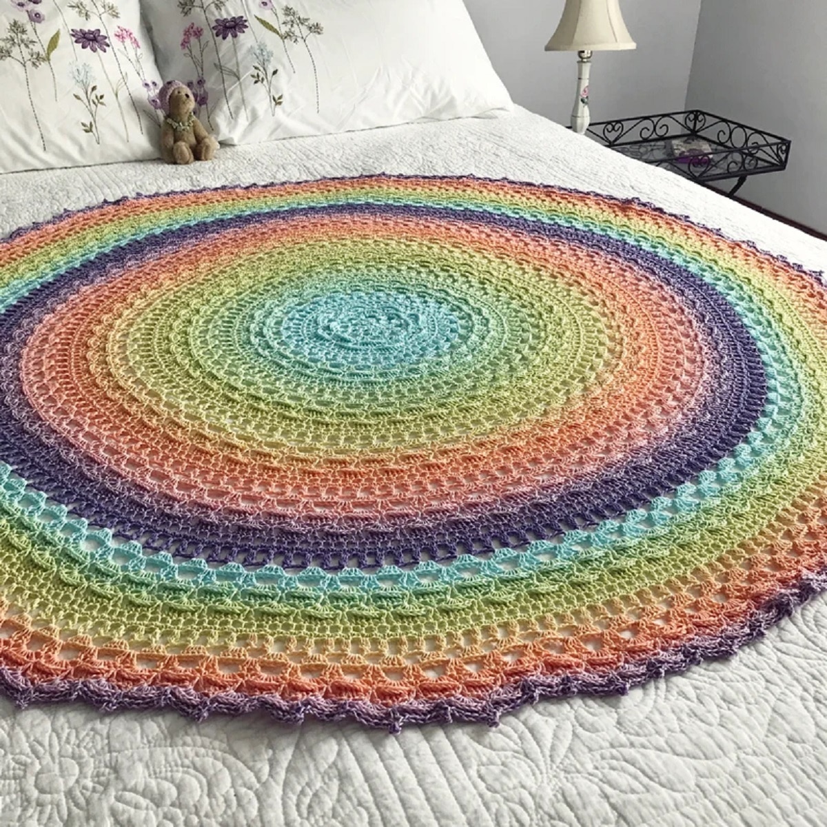 A round rainbow colored crochet mandala with circles getting smaller towards the center lying on a cream bed.