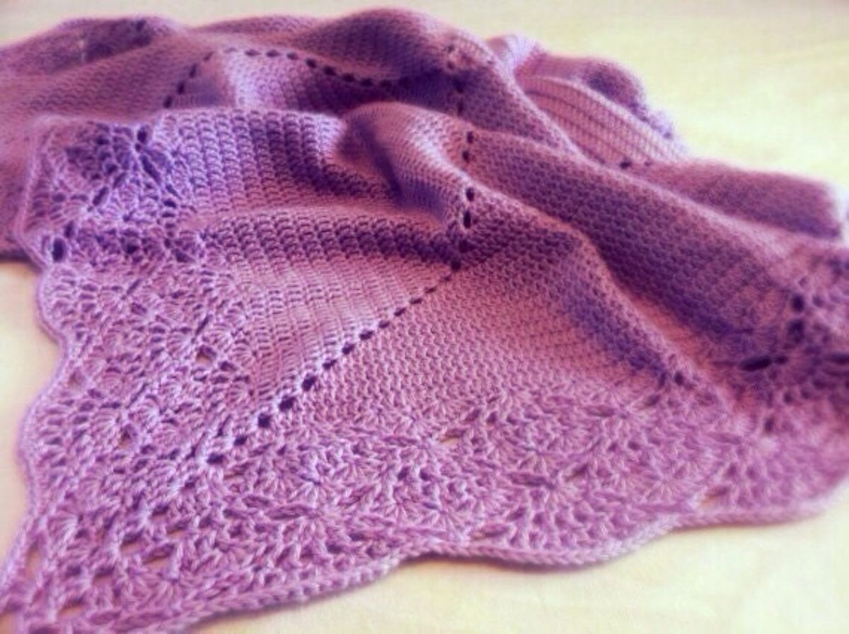 Purple crochet baby blanket with a long lace edge on all sides, bunched up on a light wooden floor.