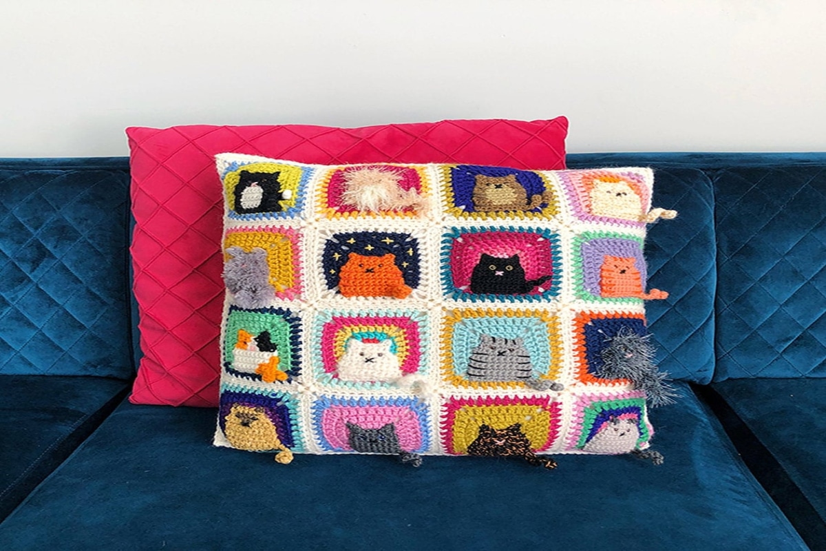 Cream crochet cushion with four rows of four squares with a different color cat stitched in each square. Cats are orange, gray, black, and white.